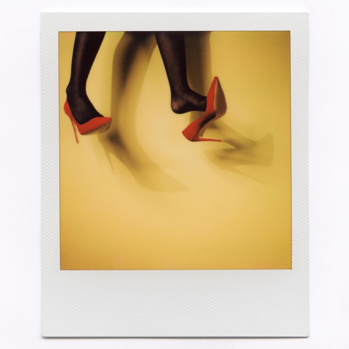 A woman's legs in black stockings and red high heels are captured mid-stride against a yellow background with blurred shadows.