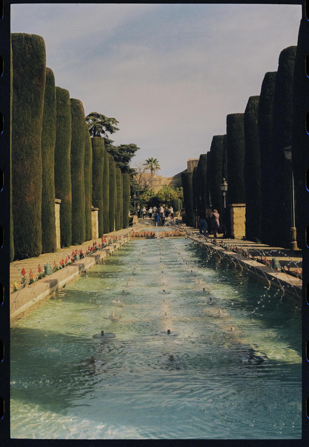 A long, narrow water feature with multiple fountains is flanked by tall, manicured hedges and a path with people in the distance.