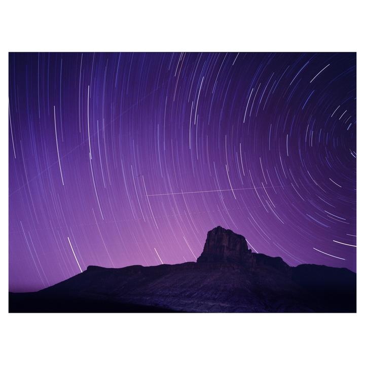 Star trails arc across a purple night sky above a silhouetted mountain.