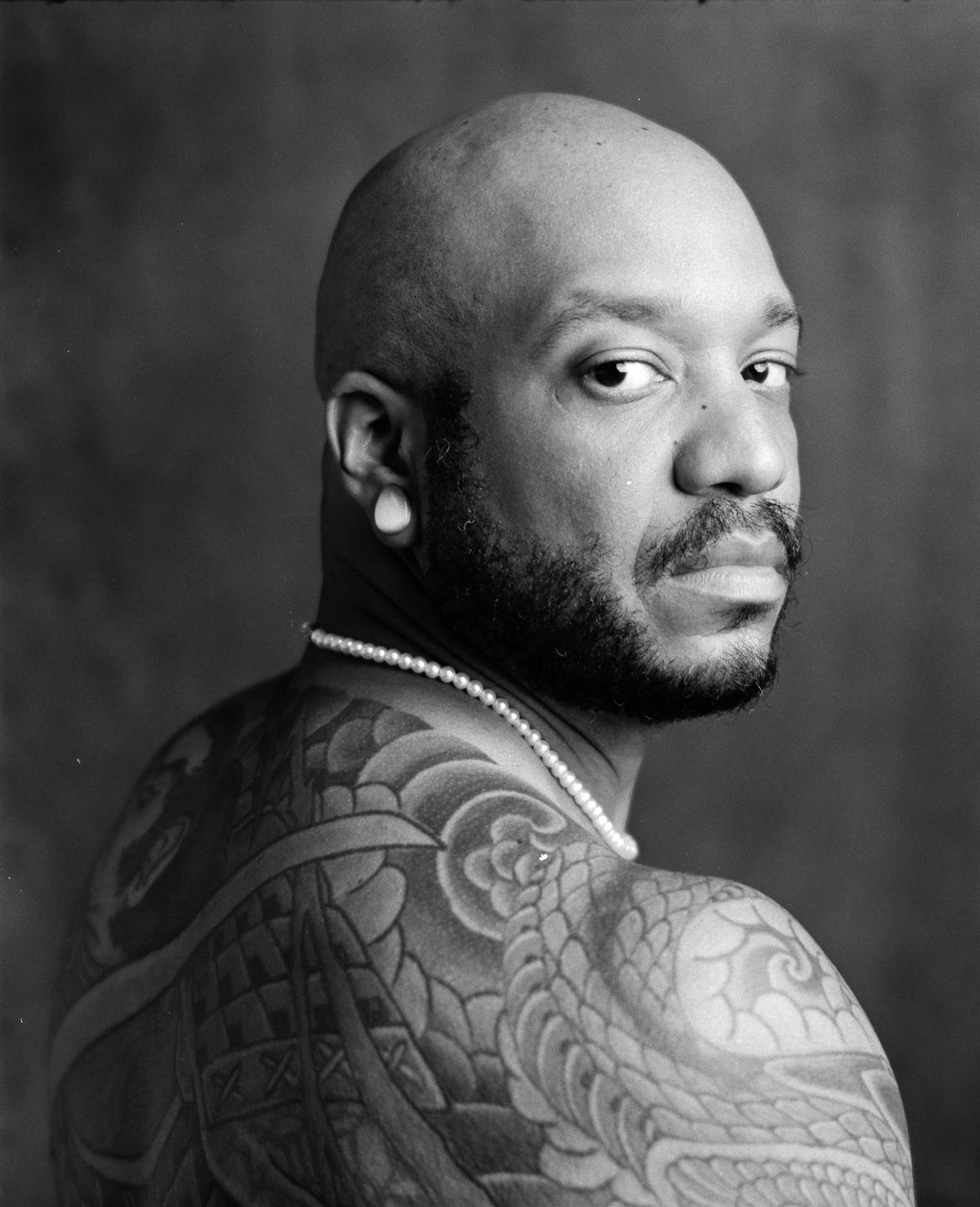 A man with a beard and a pearl necklace looks over his shoulder, revealing a tattooed back.