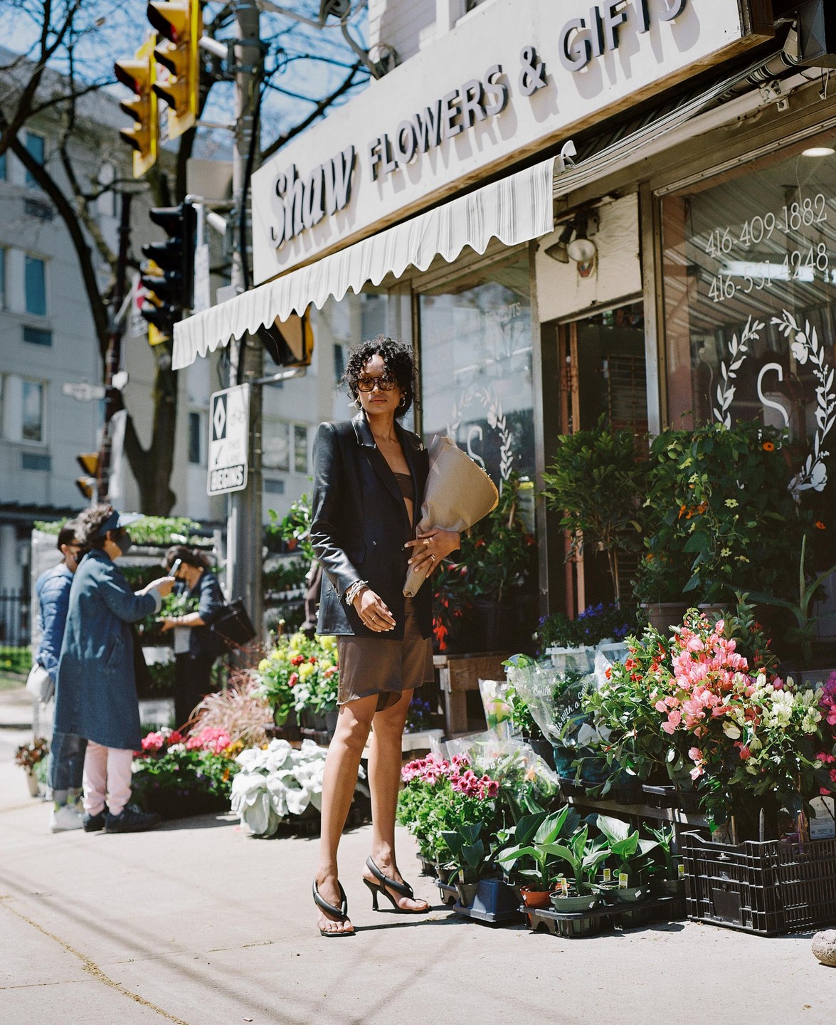 A woman stands on a sidewalk in front of a flower shop, holding a bouquet.