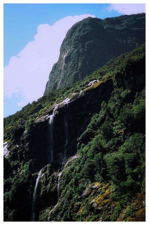 A waterfall cascades down a lush, green mountain under a blue sky with a single cloud.