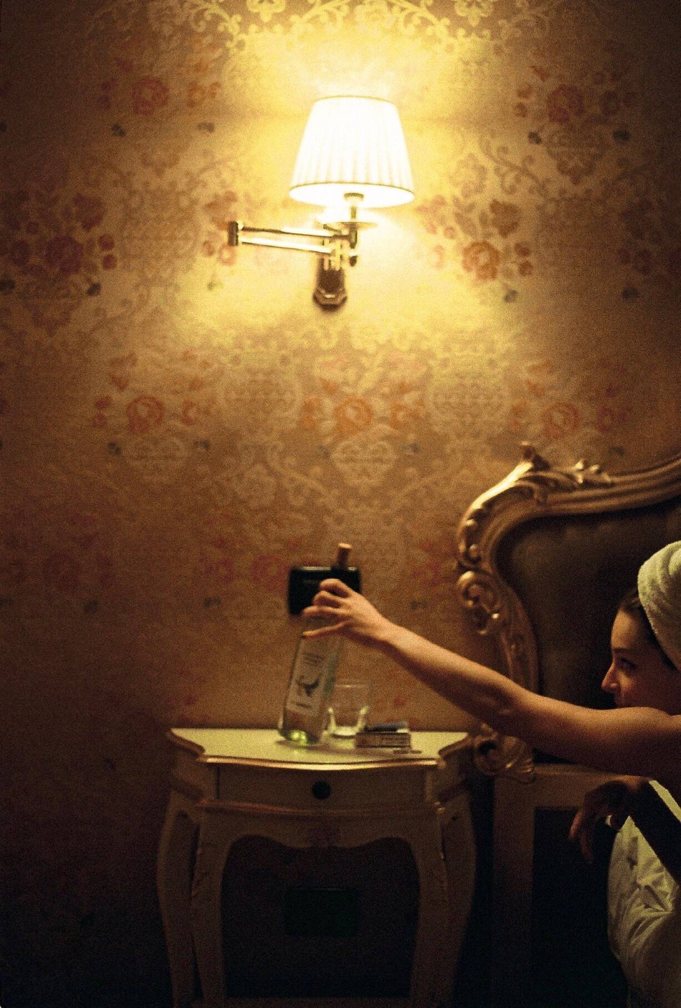 A woman with a towel on her head reaches for a bottle on a nightstand next to a bed.