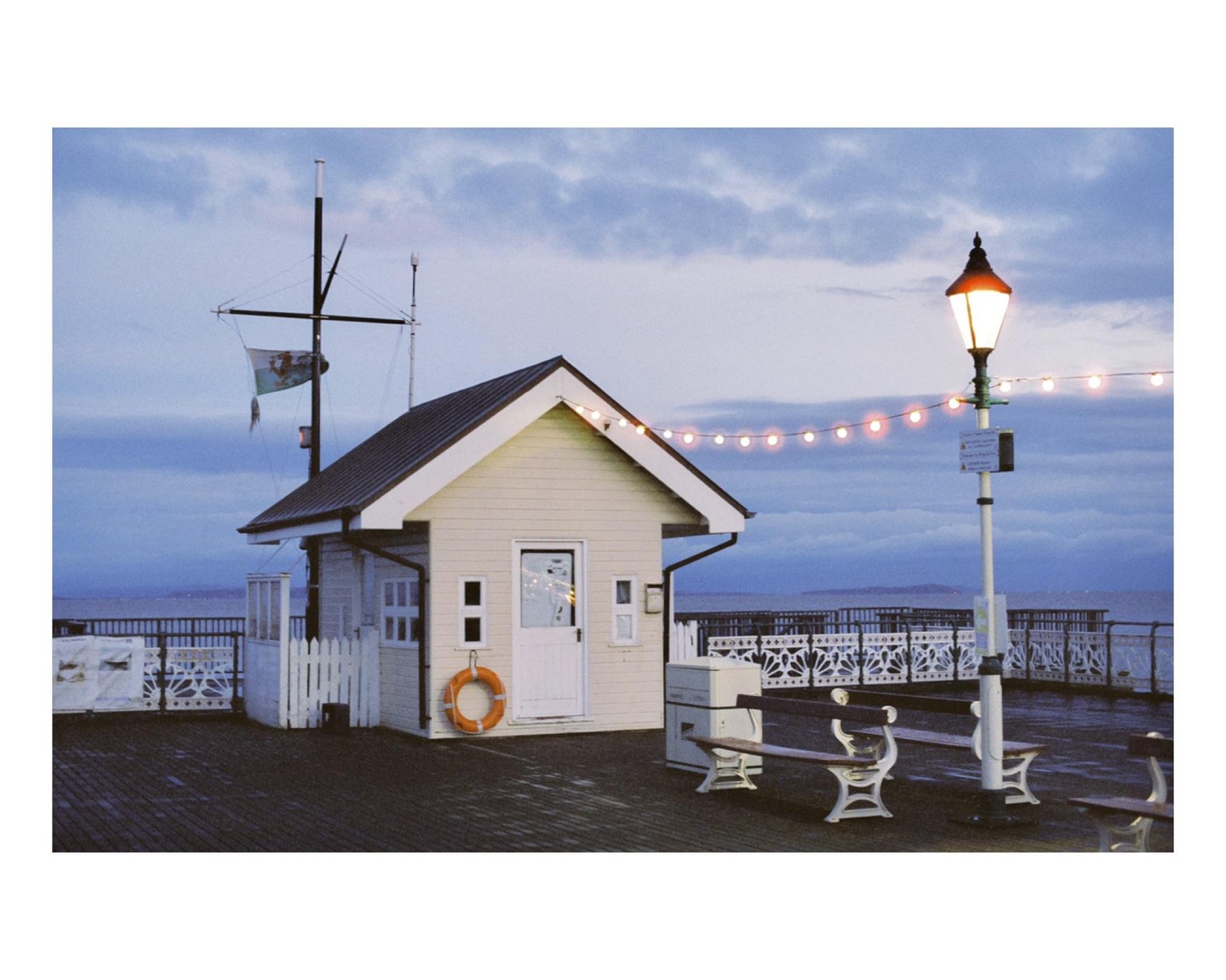 A small building with a flag pole and a street lamp with string lights stands on a pier overlooking the ocean under a cloudy sky.