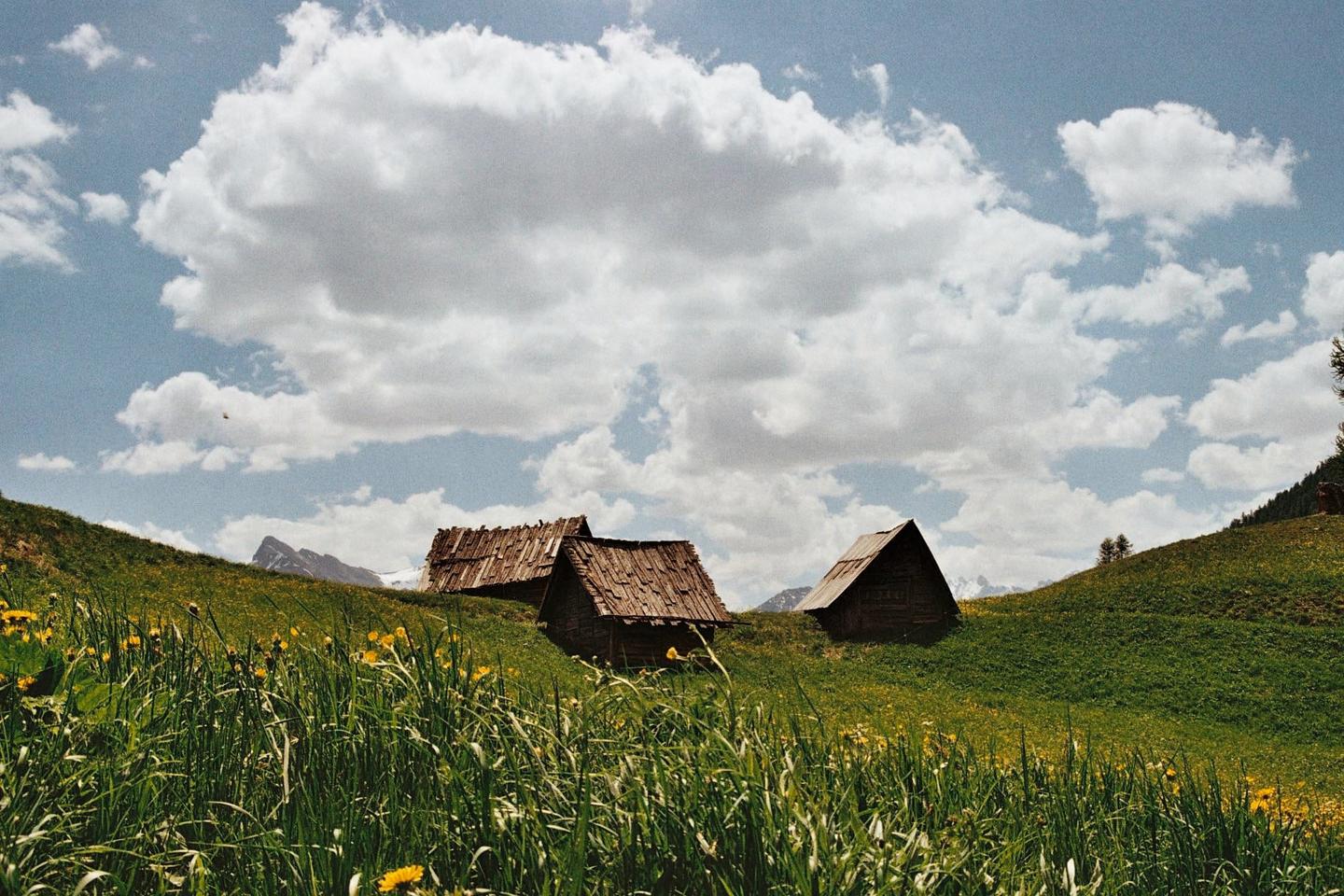 Three wooden huts with shingled roofs are nestled on a grassy hillside under a cloudy sky.