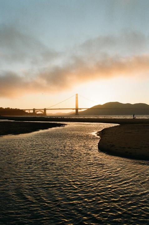 A river flows into a body of water with a suspension bridge and mountains in the background at sunset.