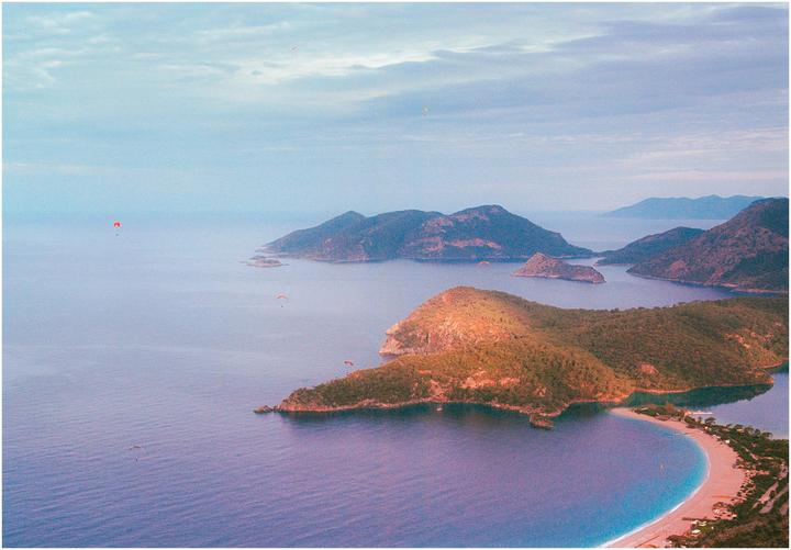 A high-angle landscape shows a blue lagoon with a sandy beach, surrounded by green hills and islands, with several paragliders in the sky.
