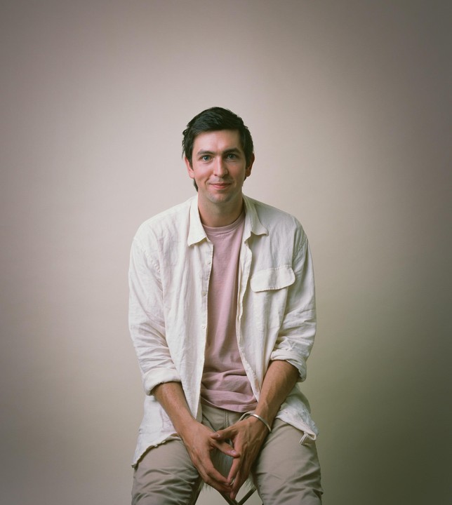 A man with dark hair and a light shirt sits on a stool with his hands clasped.