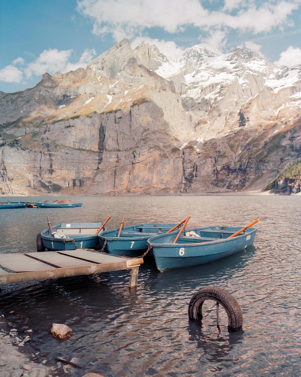 Three blue rowboats are tied to a wooden dock on a lake with snow-capped mountains in the background.