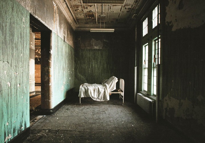 An abandoned room with peeling green paint on the walls contains a metal bed frame with white sheets and tall windows.