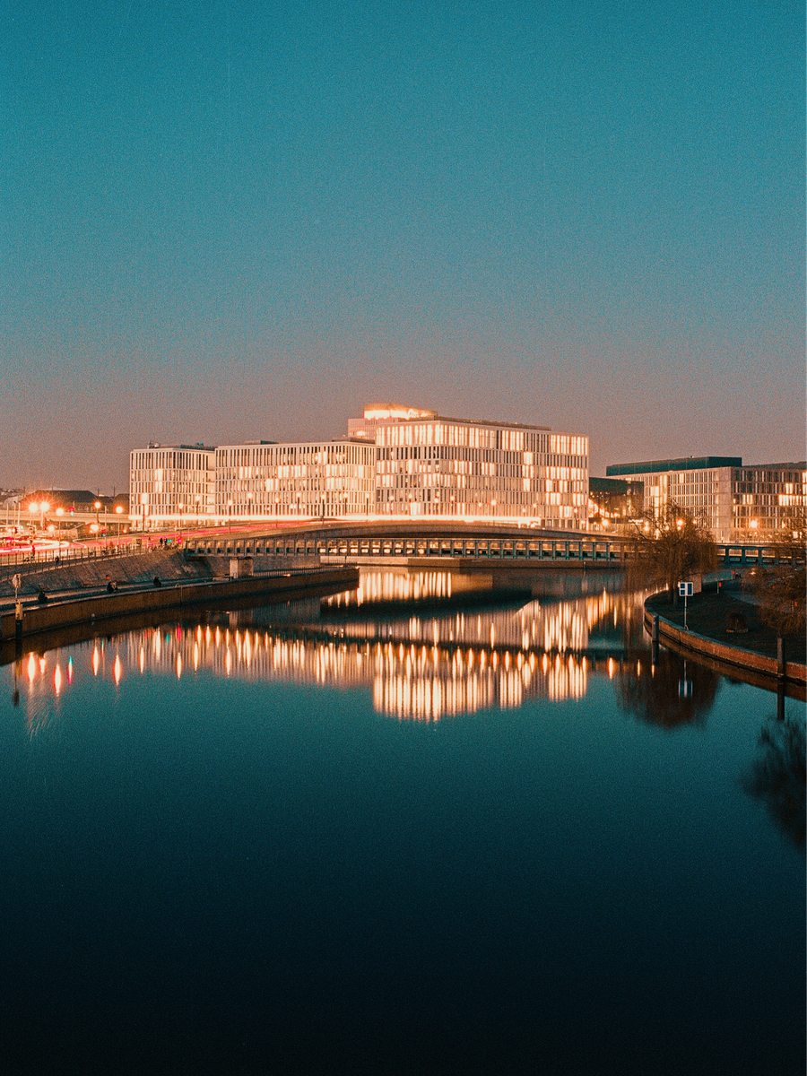 A modern building with many illuminated windows is reflected in the dark water of a river at dusk.
