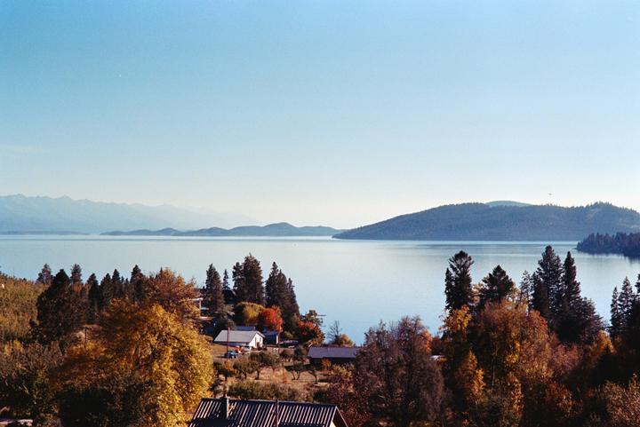 A wide shot shows a lake with mountains in the distance and houses nestled among trees in the foreground.