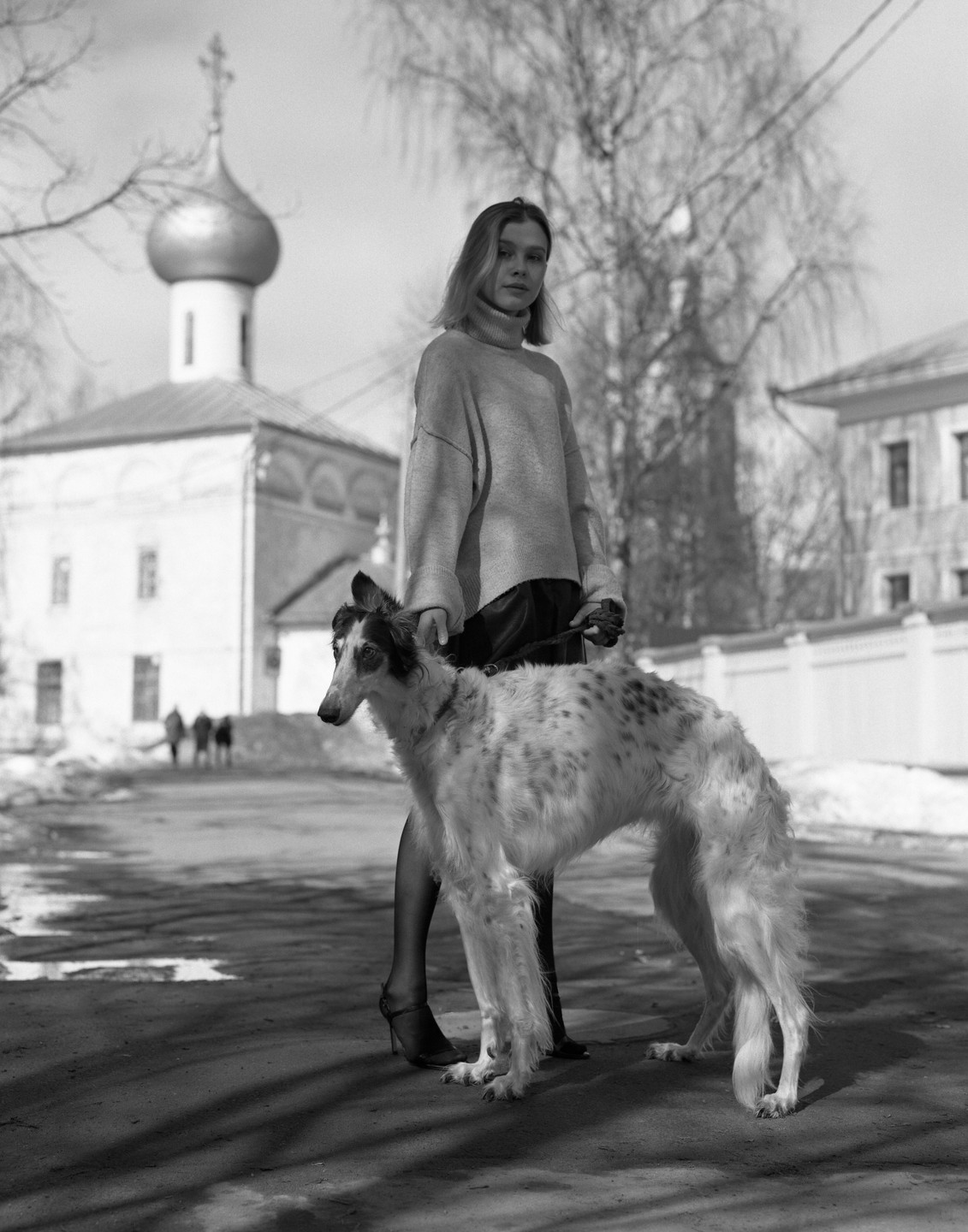 A woman stands on a street holding a borzoi on a leash, with a church in the background.