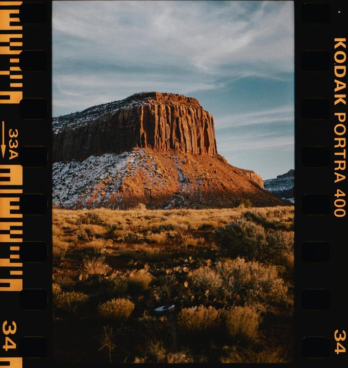 A large mesa with a flat top and steep sides rises from a desert landscape, with patches of snow on its lower slopes.