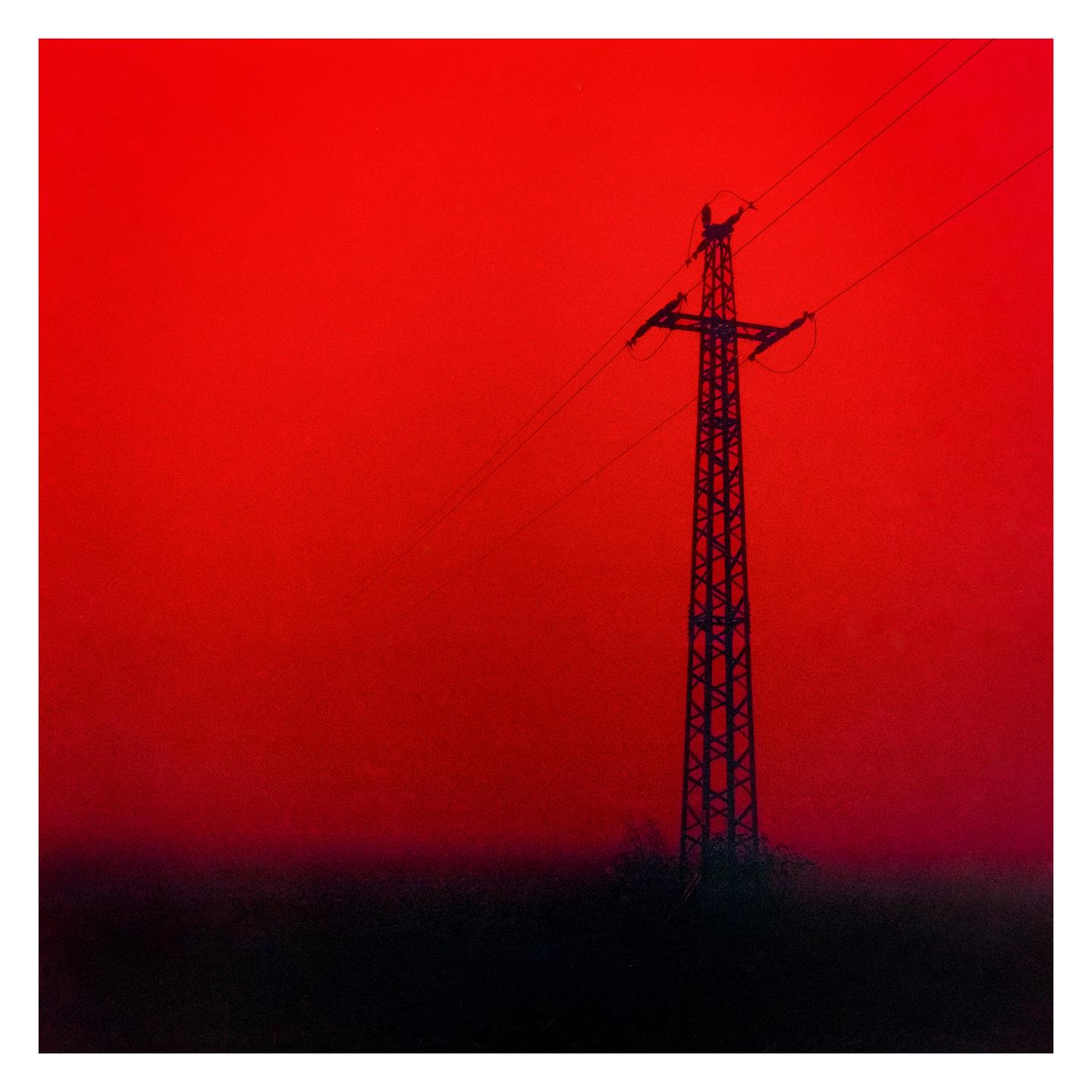 A power line tower stands silhouetted against a vibrant red sky.