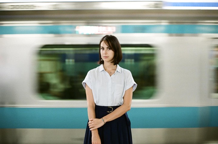A woman stands on a subway platform with a blurred train passing behind her.
