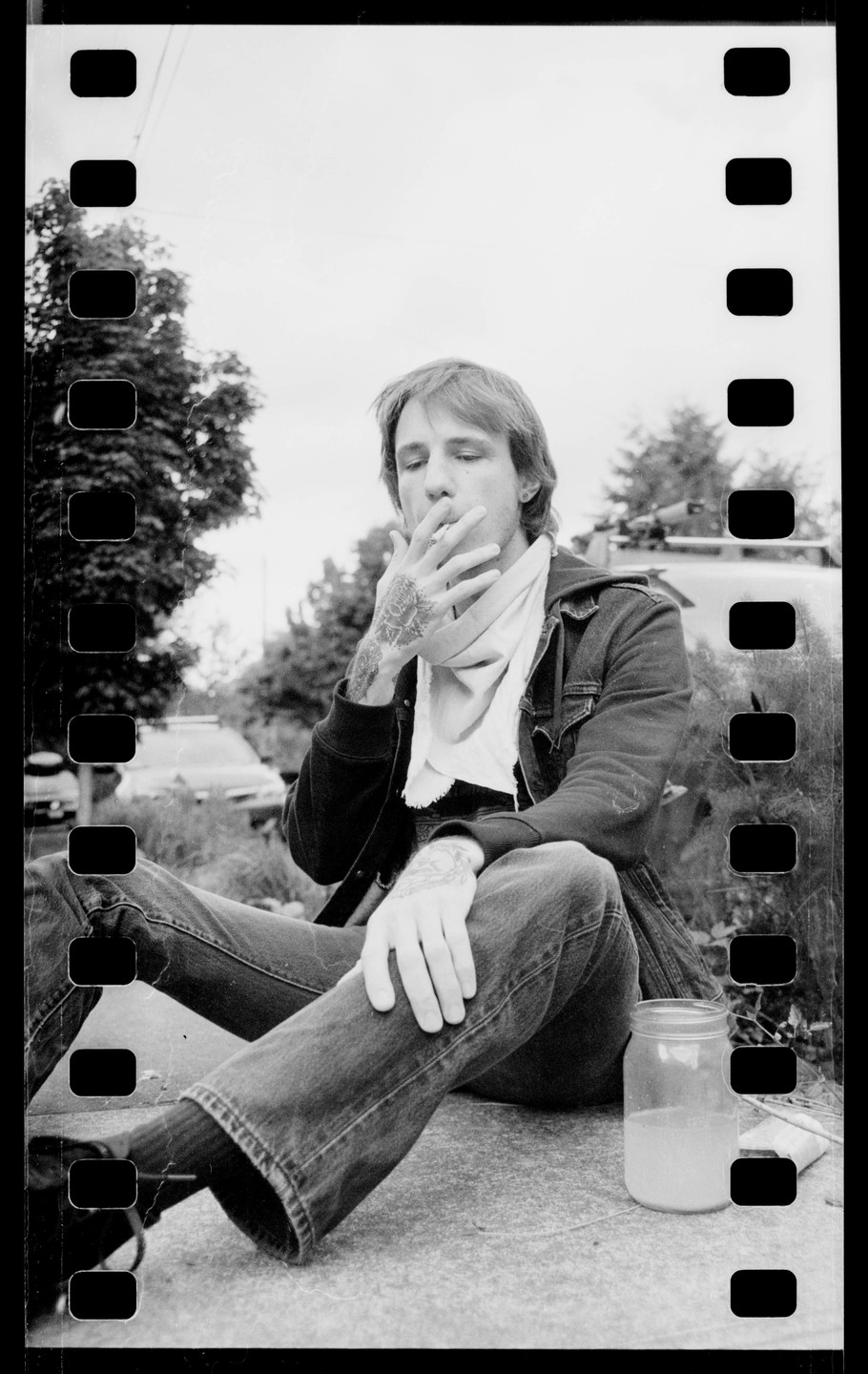 A man with tattoos on his hands sits outdoors, smoking a cigarette, with a jar of liquid beside him.