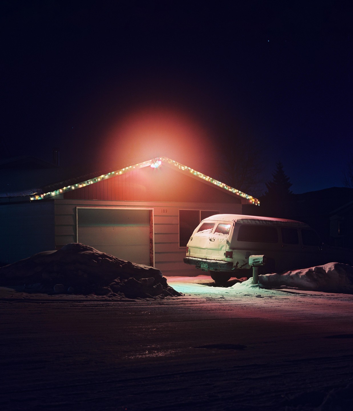 A house with Christmas lights and a van covered in snow are illuminated at night.