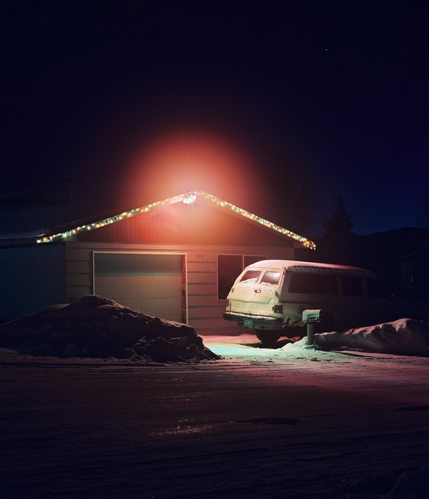 A house with Christmas lights and a van covered in snow are illuminated at night.