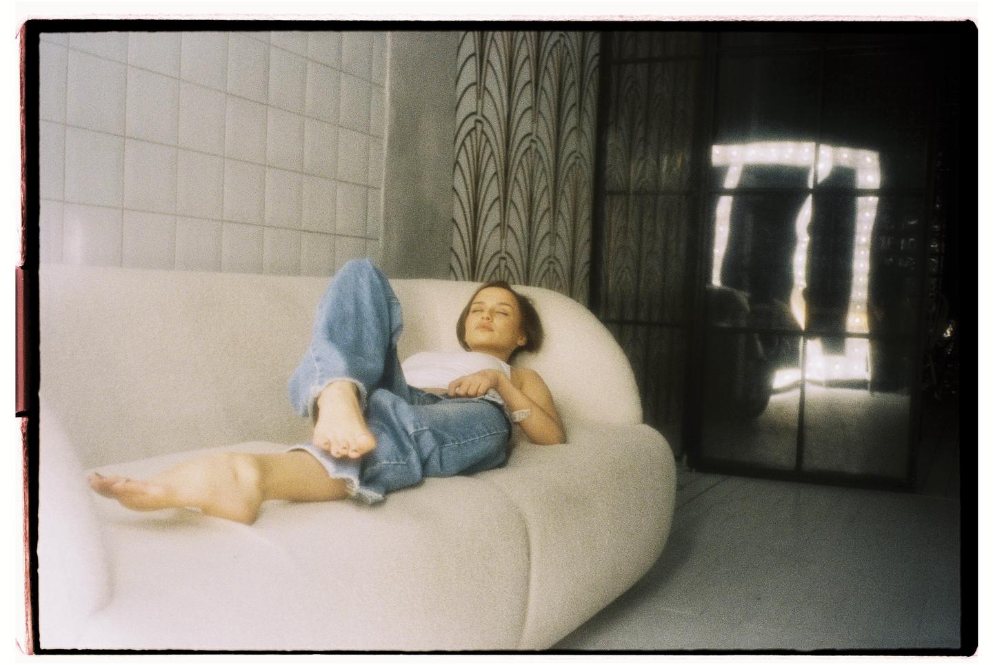 A woman in jeans and a white top is lying on a white sofa with her eyes closed.
