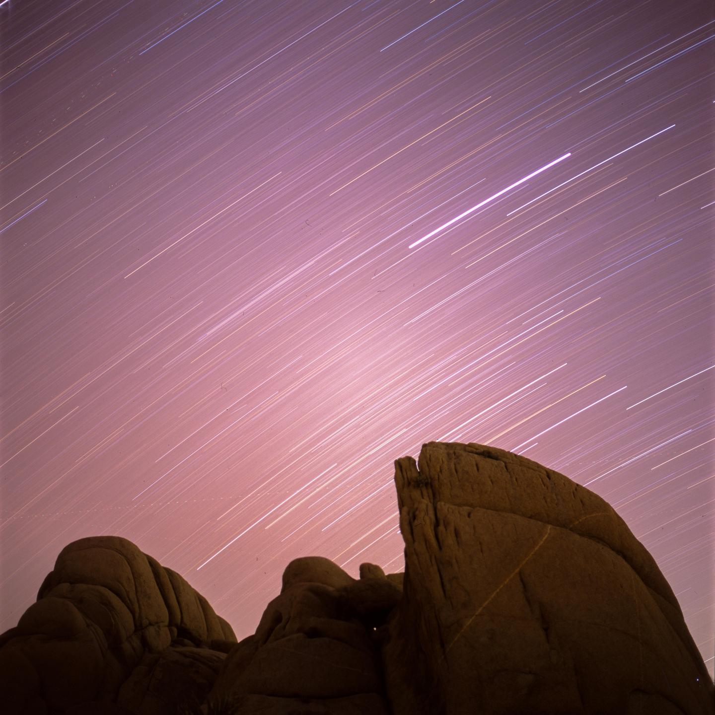 Star trails streak across a pink night sky above dark rock formations.