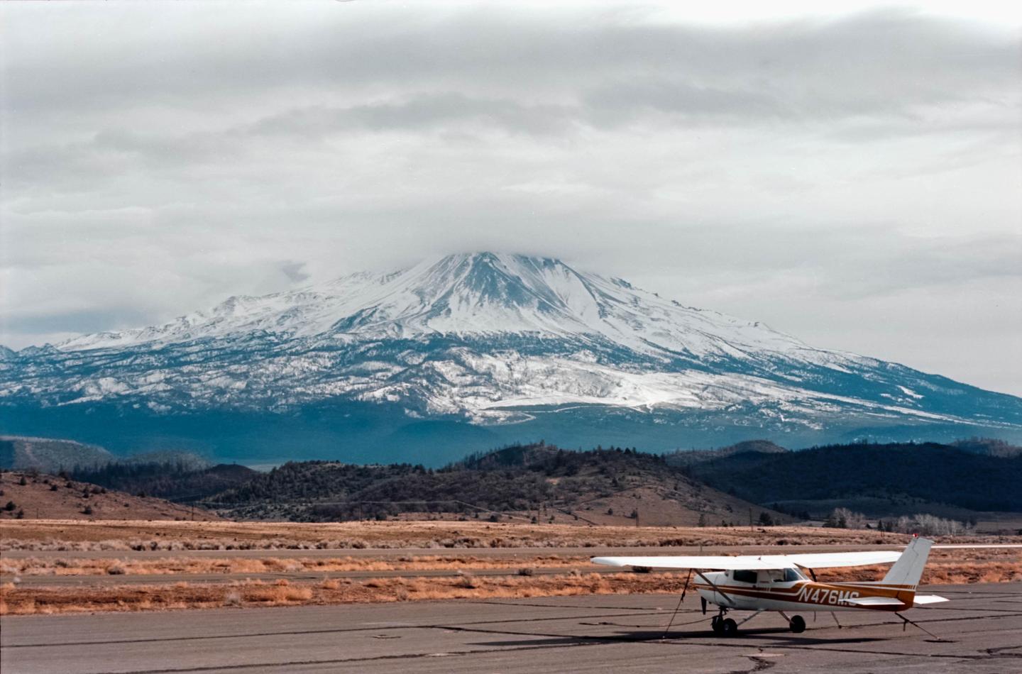 A small airplane is parked on an airfield with a large snow-capped mountain in the background under a cloudy sky.