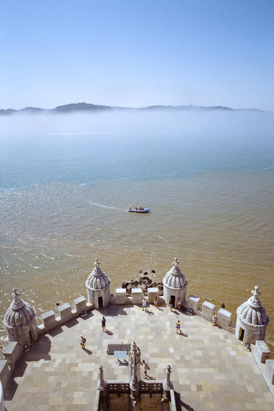 A high-angle view shows the top of a stone tower with people walking on its battlements, overlooking a body of water with a boat and a hazy distant shoreline.
