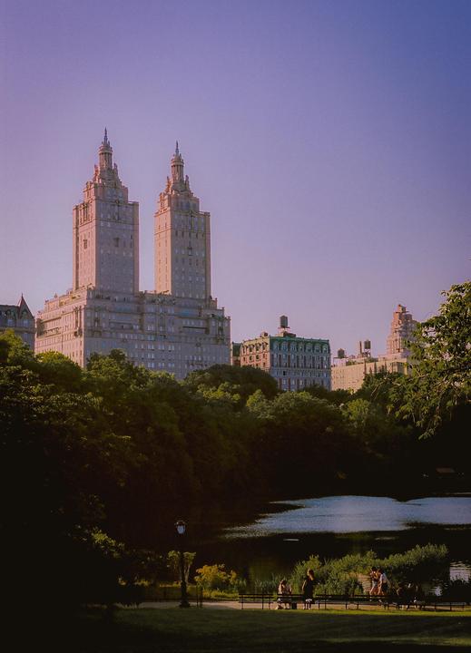 Two tall buildings with ornate spires rise above a treeline, overlooking a body of water where people are gathered on benches.