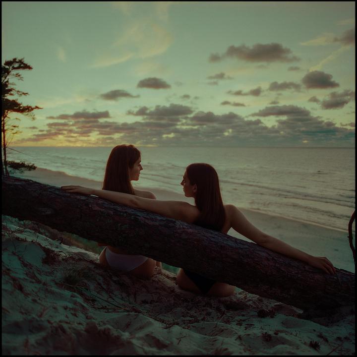 Two women sit on a sandy beach next to a fallen tree trunk, looking at each other with the ocean and a cloudy sky in the background at sunset.