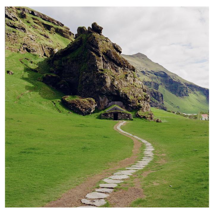 A winding stone path leads to a small turf house built into the base of a large, moss-covered rock formation in a green, mountainous landscape.