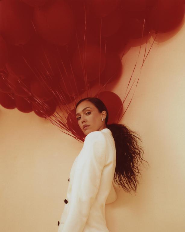 A woman in a white blazer stands in front of a wall with red balloons.