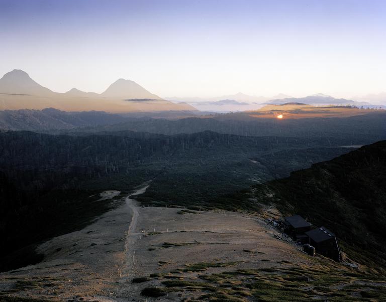 A double exposure shows a sunset over a forest and mountains, with a path leading to a building on a ridge.