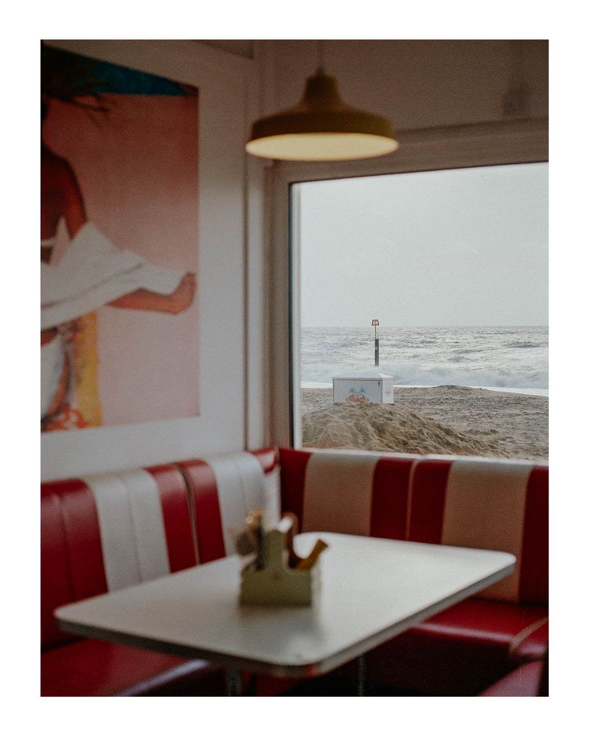 A diner booth with red and white striped seating and a table is visible through a window overlooking a beach and ocean.