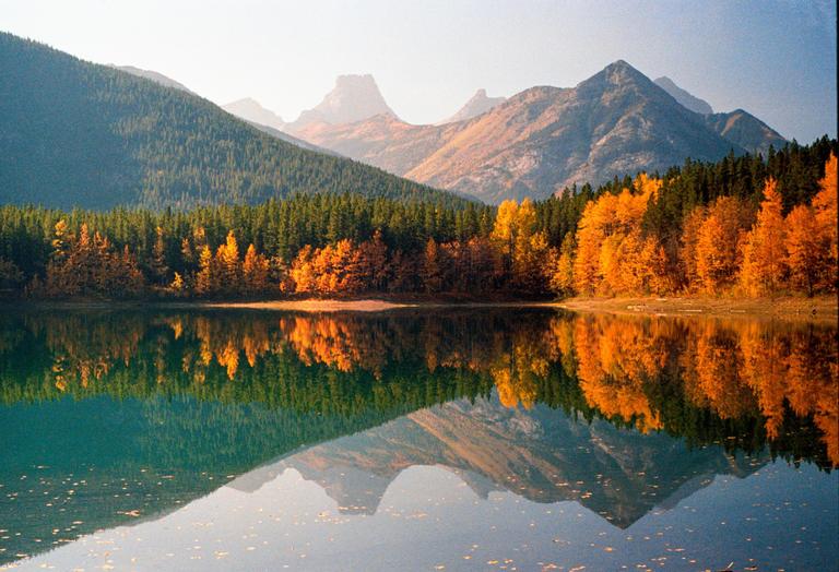 A lake reflects a forest with autumn foliage and mountains under a clear sky.