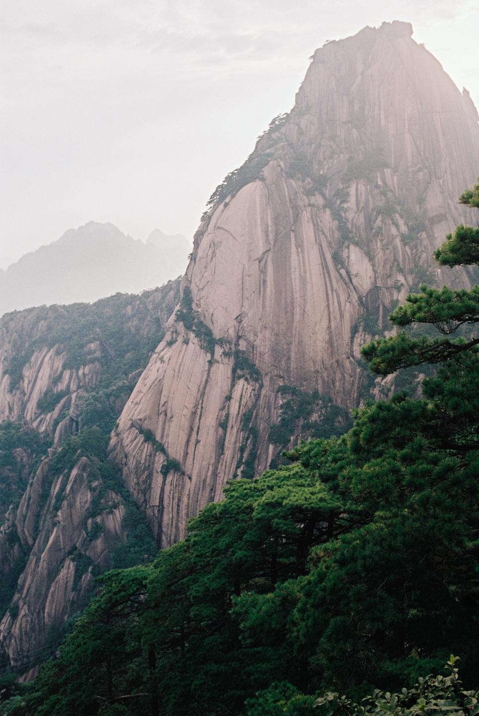 A large, rocky mountain with green trees in the foreground rises into a hazy sky.