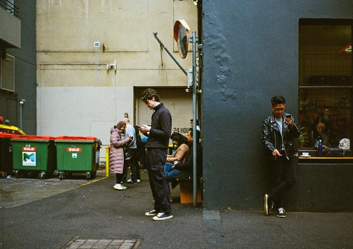A man in a leather jacket leans against a dark wall while looking at his phone, with other people and green bins visible in the background.