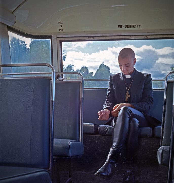 A woman with a shaved head wearing a clerical collar and a cross necklace sits on a bus.