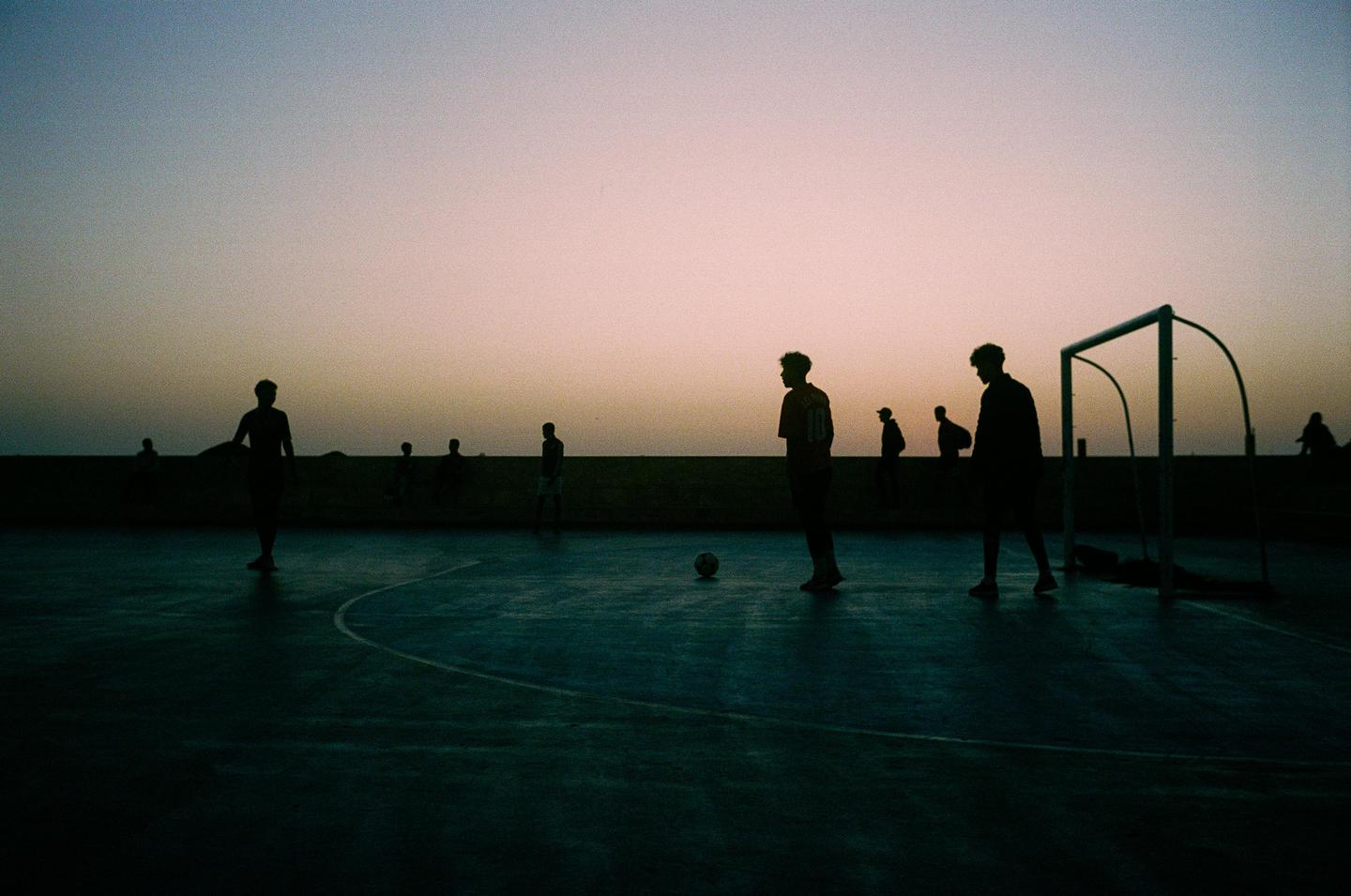 Silhouetted men play soccer on a court at dusk.