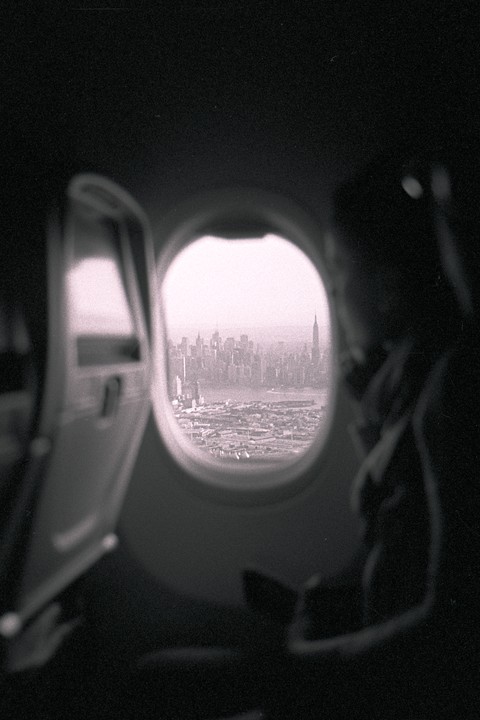 A hazy view of the New York City skyline with the Empire State Building visible through an airplane window, with a person's silhouette on the right.