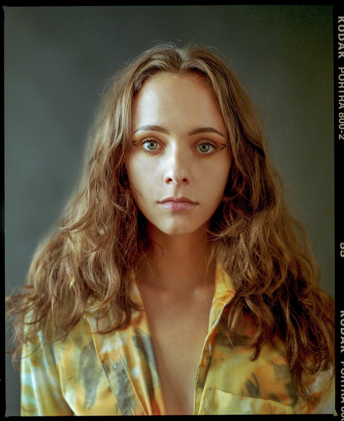 A woman with long wavy hair and a patterned shirt looks directly at the viewer.