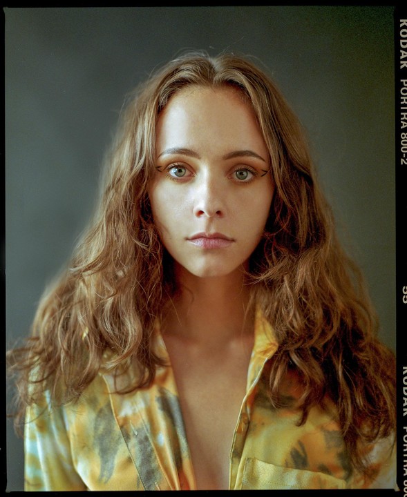 A woman with long wavy hair and a patterned shirt looks directly at the viewer.