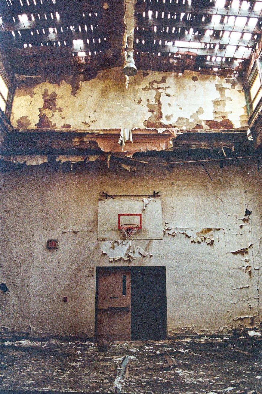 A basketball hoop is mounted on a wall with peeling paint in an abandoned school.