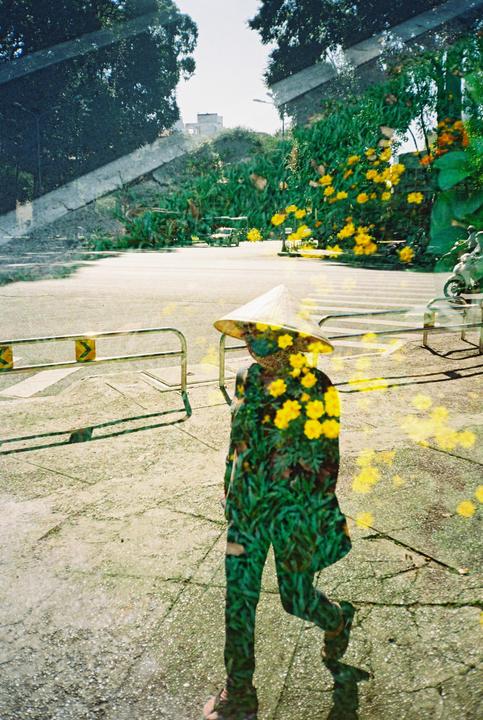 A double exposure shows a person wearing a conical hat walking on a street, overlaid with an image of yellow flowers and green foliage.
