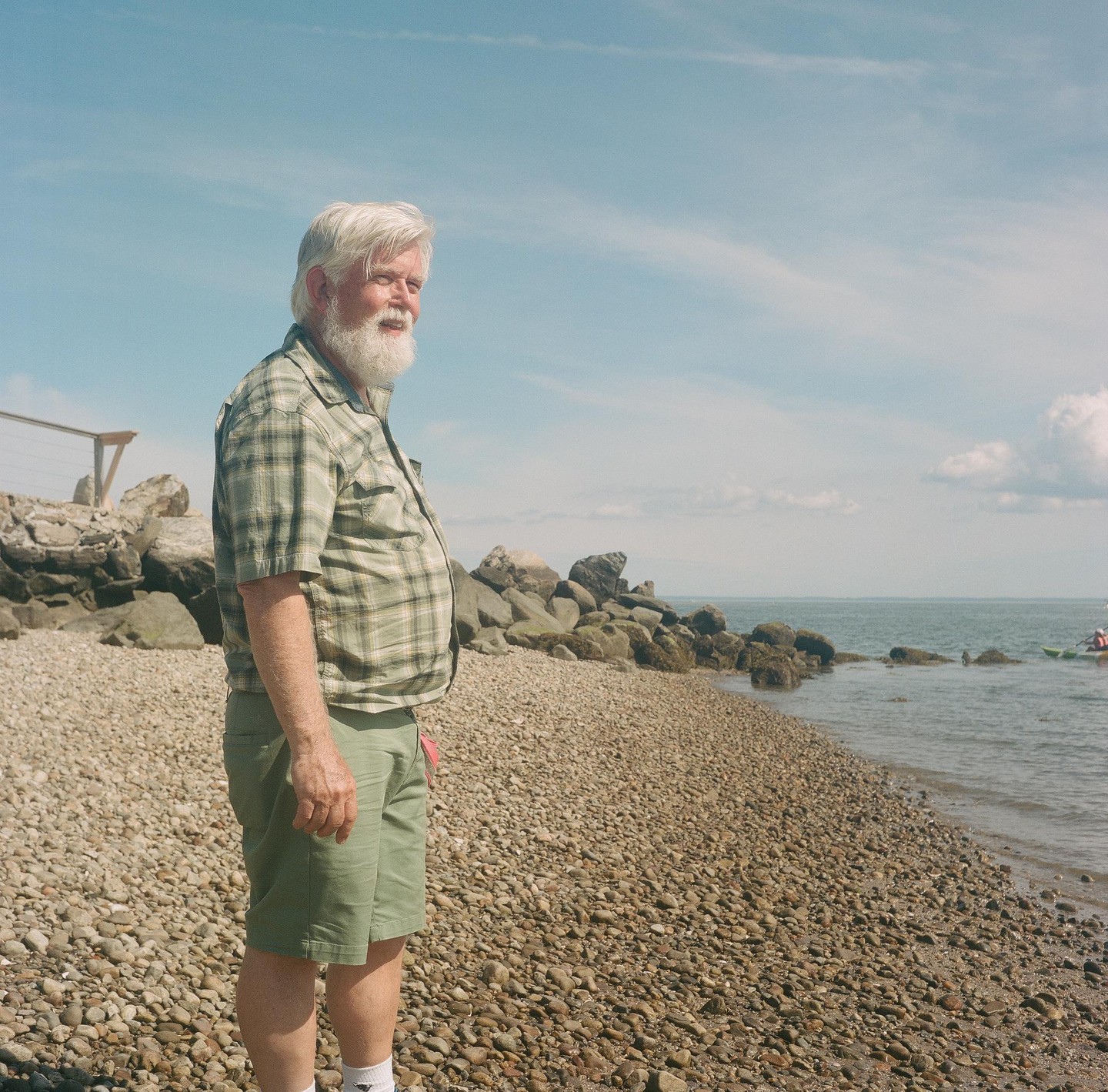 A man with a white beard stands on a rocky beach looking out at the water.
