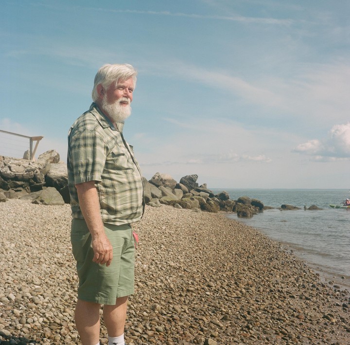 A man with a white beard stands on a rocky beach looking out at the water.