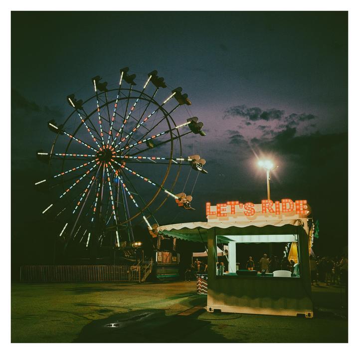 A brightly lit Ferris wheel and a "Let's Ride" booth stand out against a dark, cloudy night sky.