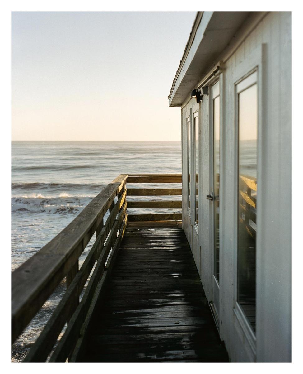 A wooden pier with a building on one side extends over the ocean at sunrise.