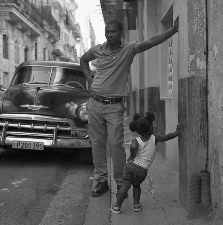 A man and a child stand on a sidewalk next to a vintage car in Havana.