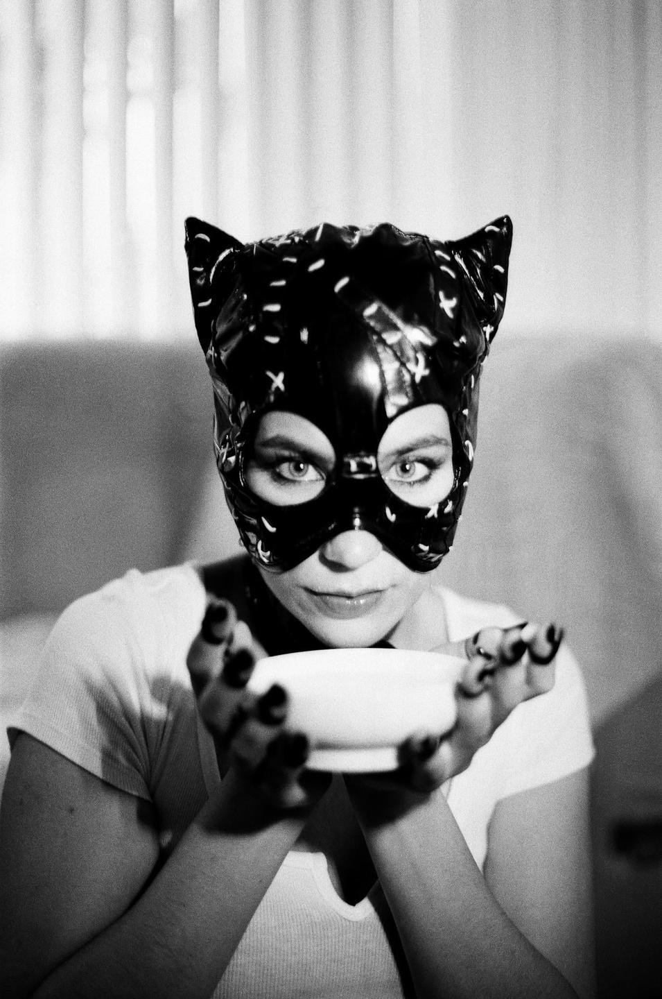 A woman wearing a cat mask holds a small bowl with both hands.