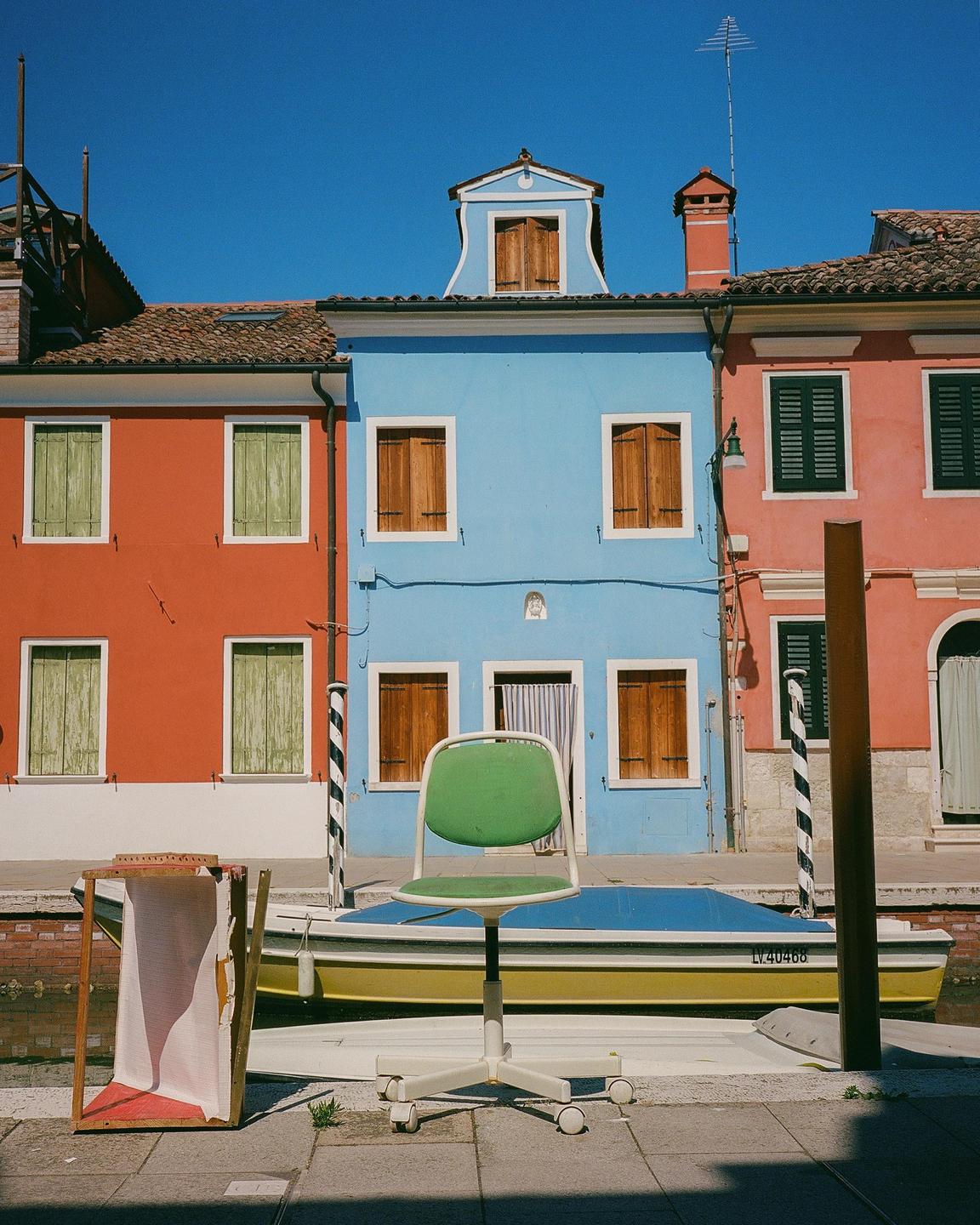 A green office chair sits on a paved surface in front of a canal with a boat and colorful buildings.